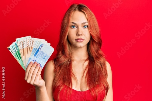 Young beautiful redhead woman holding south korean won banknotes thinking attitude and sober expression looking self confident
