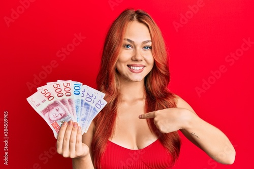 Young beautiful redhead woman holding swedish krona banknotes pointing finger to one self smiling happy and proud
