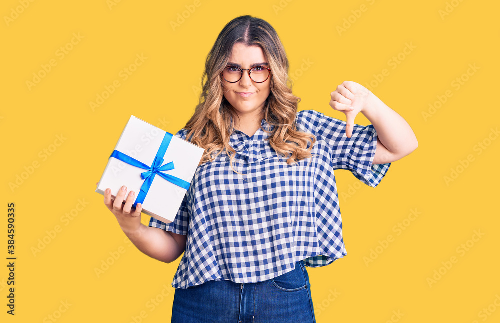 Young caucasian woman holding gift with angry face, negative sign showing dislike with thumbs down, rejection concept Young caucasian woman holding gift with angry face, negative sign showing dislike with thumbs down, rejection concept