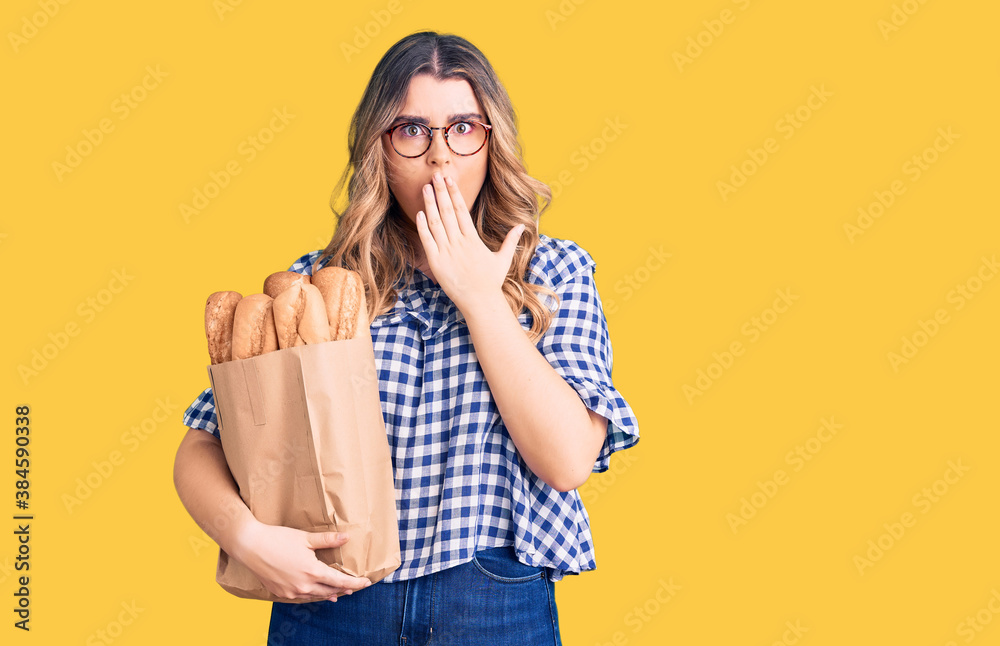 Young caucasian woman holding paper bag with bread covering mouth with hand, shocked and afraid for mistake. surprised expression Young caucasian woman holding paper bag with bread covering mouth with hand, shocked and afraid for mistake. surprised expression