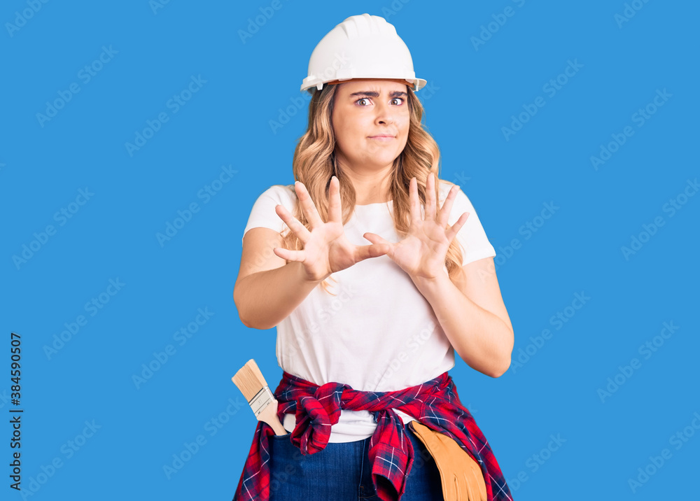 Young caucasian woman wearing security helmet afraid and terrified with fear expression stop gesture with hands, shouting in shock. panic concept. Young caucasian woman wearing security helmet afraid and terrified with fear expression stop gesture with hands, shouting in shock. panic concept.