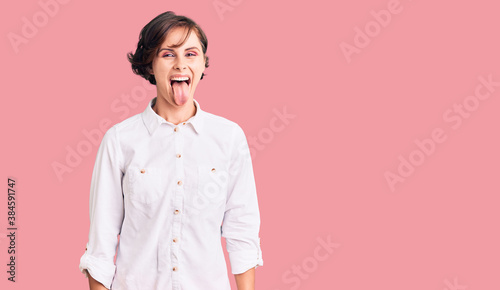 Beautiful young woman with short hair wearing elegant white shirt sticking tongue out happy with funny expression. emotion concept.