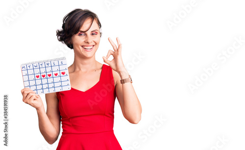 Beautiful young woman with short hair holding heart calendar doing ok sign with fingers  smiling friendly gesturing excellent symbol