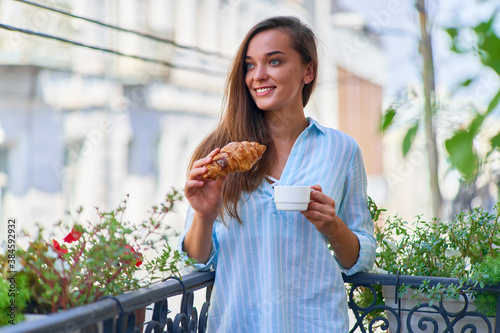 Portrait of a beautiful happy cute joyful smiling romantic woman with coffee cup and fresh baked croissant for french breakfast in the morning on a balcony photo
