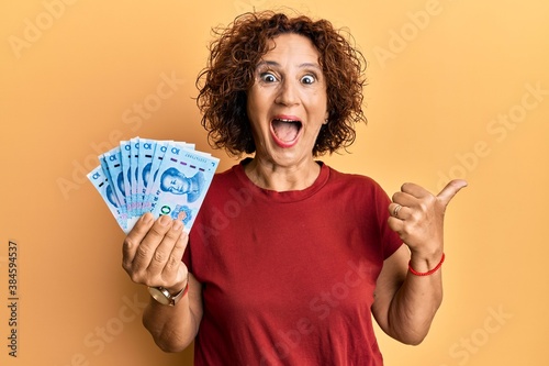 Beautiful middle age mature woman holding 10 yuan chinese banknotes pointing thumb up to the side smiling happy with open mouth
