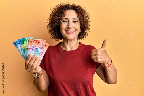 Beautiful middle age mature woman holding swiss franc banknotes smiling happy and positive, thumb up doing excellent and approval sign