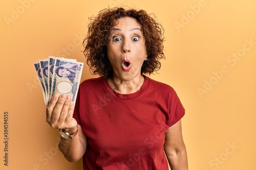Beautiful middle age mature woman holding 5000 japanese yen banknotes scared and amazed with open mouth for surprise, disbelief face