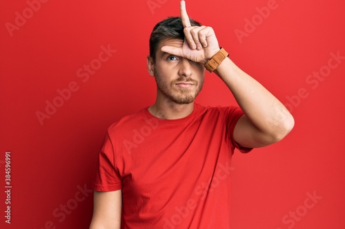 Handsome caucasian man wearing casual red tshirt making fun of people with fingers on forehead doing loser gesture mocking and insulting.