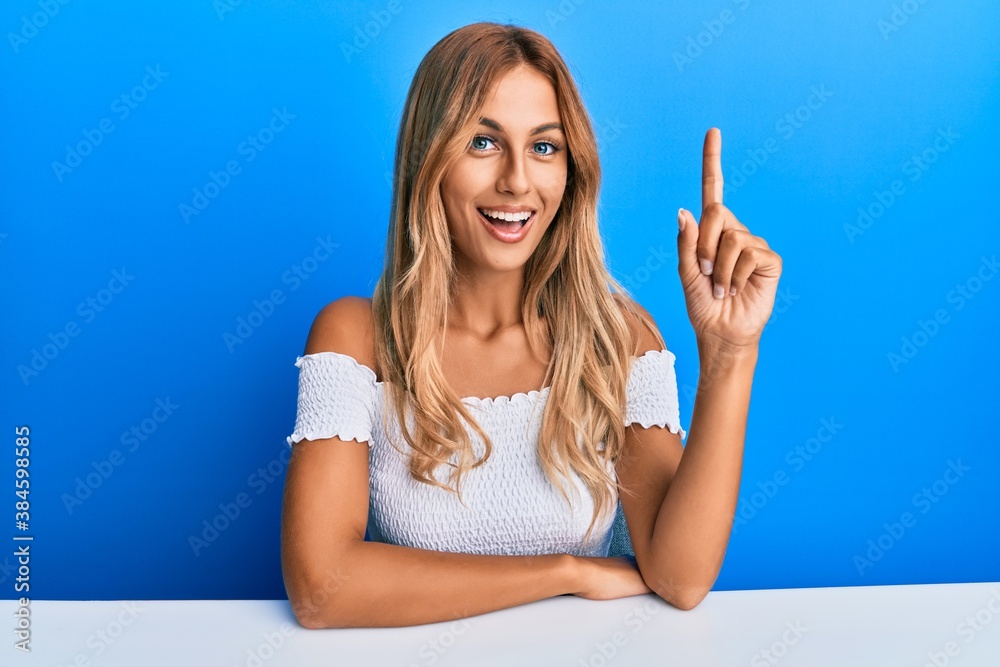 Beautiful blonde young woman wearing casual clothes sitting on the table smiling with an idea or question pointing finger up with happy face, number one Beautiful blonde young woman wearing casual clothes sitting on the table smiling with an idea or question pointing finger up with happy face, number one