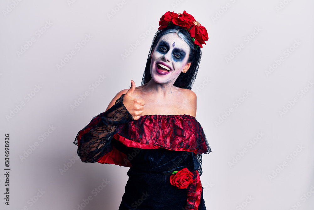 Young woman wearing mexican day of the dead makeup doing happy thumbs up gesture with hand. approving expression looking at the camera showing success. Young woman wearing mexican day of the dead makeup doing happy thumbs up gesture with hand. approving expression looking at the camera showing success.
