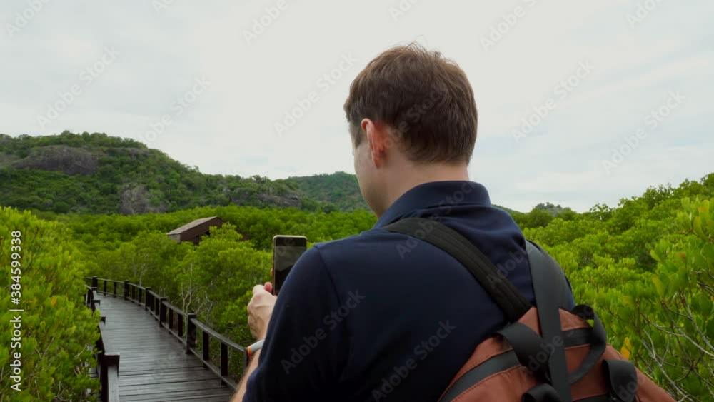 Young Tourist Man with Backpack Makes Video on Smartphone in Green Sunny Forest. Back View. Traveler Shooting Clip for Stories to Social Media by Phone Standing on Wooden Boardwalk in Woods. Young Tourist Man with Backpack Makes Video on Smartphone in Green Sunny Forest. Back View. Traveler Shooting Clip for Stories to Social Media by Phone Standing on Wooden Boardwalk in Woods.