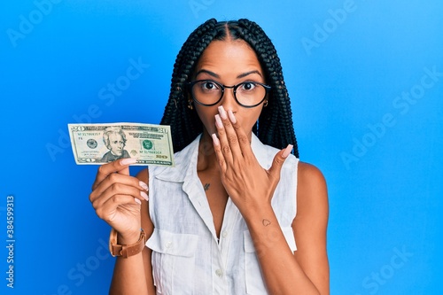 Beautiful hispanic woman holding 20 dollars banknote covering mouth with hand, shocked and afraid for mistake. surprised expression