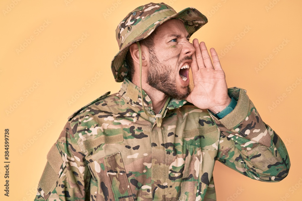 Young caucasian man wearing camouflage army uniform shouting and screaming loud to side with hand on mouth. communication concept.