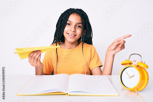 Young african american girl child with braids holding paper airplane while studying smiling happy pointing with hand and finger to the side photo