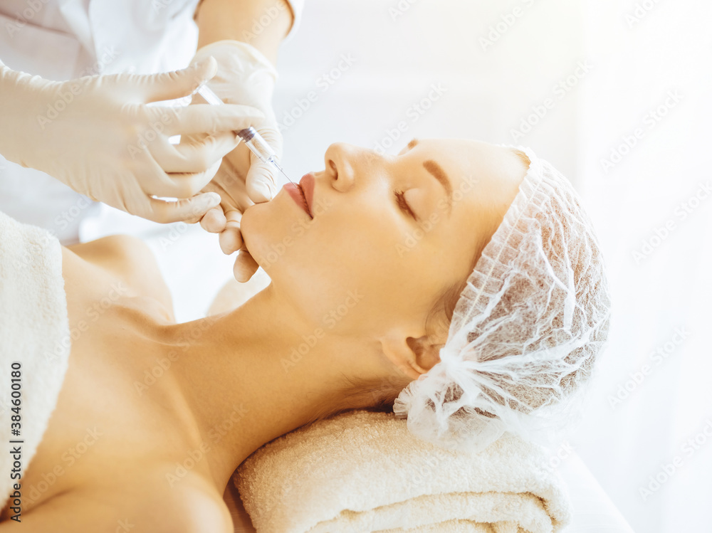 Fototapeta premium Beautiful woman receiving beauty injections with closed eyes in sunny medical center. Beautician doctor hands doing beauty procedure to female face with syringe
