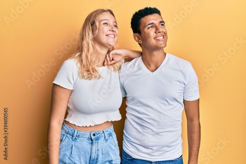 Young interracial couple wearing casual white tshirt looking away to side with smile on face, natural expression. laughing confident.