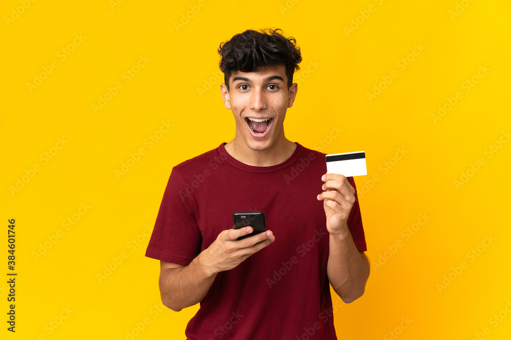 Young Argentinian man isolated on yellow background buying with the mobile and holding a credit card with surprised expression Young Argentinian man isolated on yellow background buying with the mobile and holding a credit card with surprised expression