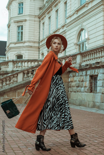 Fashionable woman wearing long orange trench coat, hat, zebra print midi skirt, cowboy ankle boots, with green bag, walking in street of European city
