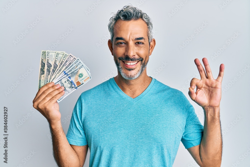 Middle age grey-haired man holding dollars doing ok sign with fingers, smiling friendly gesturing excellent symbol Middle age grey-haired man holding dollars doing ok sign with fingers, smiling friendly gesturing excellent symbol