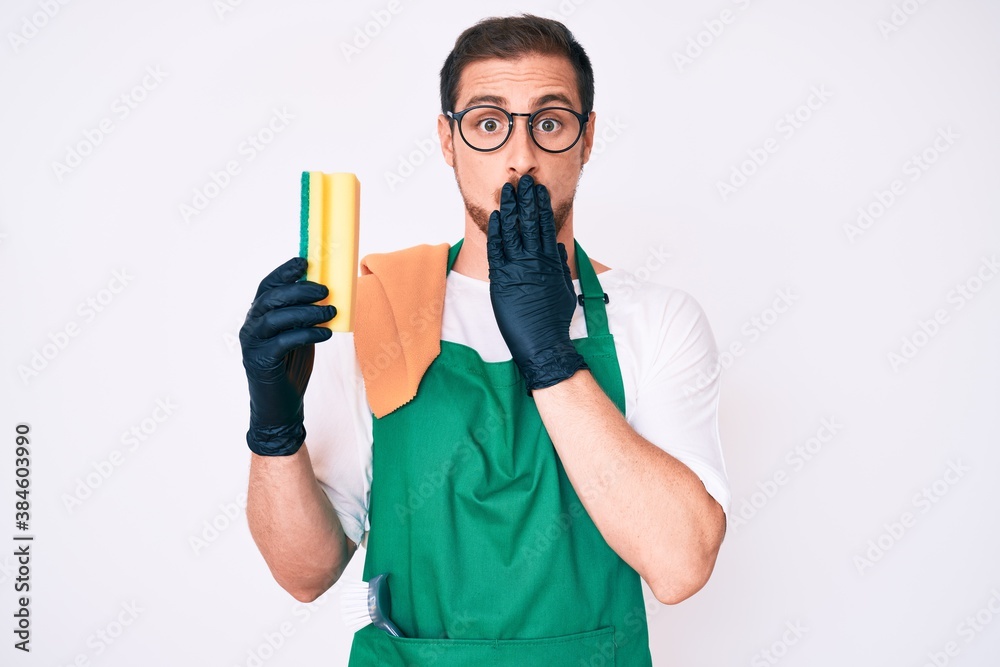 Young handsome man wearing apron holding scourer covering mouth with hand, shocked and afraid for mistake. surprised expression Young handsome man wearing apron holding scourer covering mouth with hand, shocked and afraid for mistake. surprised expression