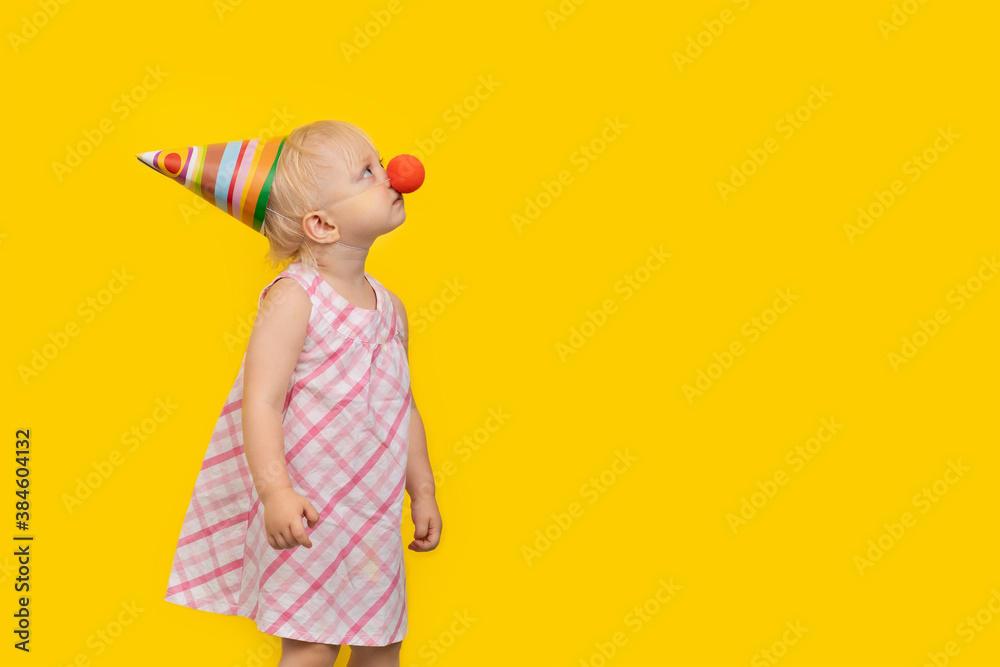 Toddler girl wearing party hat and red clown nose and looks up. Portrait on yellow background. Copy space. Toddler girl wearing party hat and red clown nose and looks up. Portrait on yellow background. Copy space.