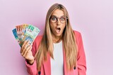 Young blonde woman wearing business style holding australian dollars scared and amazed with open mouth for surprise, disbelief face