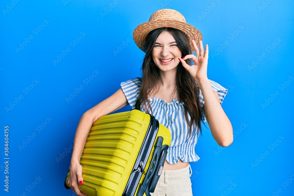 Young beautiful caucasian girl wearing summer dress and holding cabin bag doing ok sign with fingers, smiling friendly gesturing excellent symbol