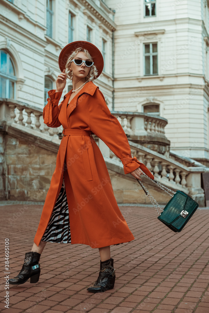 Outdoor full body autumn portrait of elegant model wearing long orange trench coat, hat, sunglasses, cowboy ankle boots, with green croco textured bag, posing in street of European city