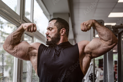 strong young athlete man with beard wearing black tank top showing big double biceps muscle in sport gym with window