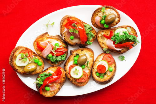 Assorted crostini canapes with cottage cheese, baked capsicum, cucumber slices, bacon, tomatoes, green peas on a festive red background. Top view,