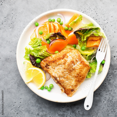 Fried fillet of sea white cod fish with juicy lettuce, capsicum, lemon, green peas on a large white dish on a gray background. Healthy balanced food. Top view,