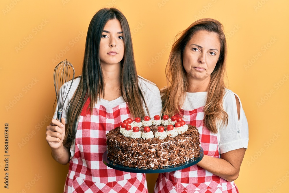 Hispanic family of mother and daughter wearing baker apron holding homemade cake relaxed with serious expression on face. simple and natural looking at the camera.