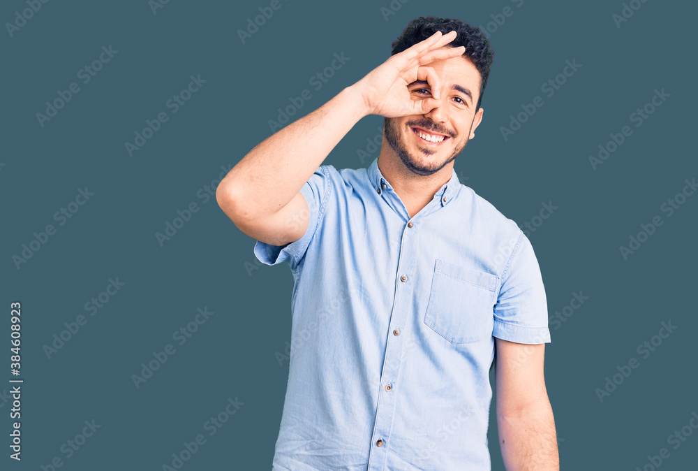 Young hispanic man wearing casual clothes smiling happy doing ok sign with hand on eye looking through fingers Young hispanic man wearing casual clothes smiling happy doing ok sign with hand on eye looking through fingers
