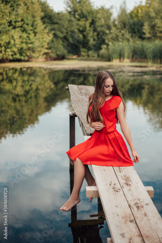 Vertical full length portrait of young woman sitting on the wooden pier near the river or lake touching clothes, dressed in red dress, looking away, copy space and nature blur background.
