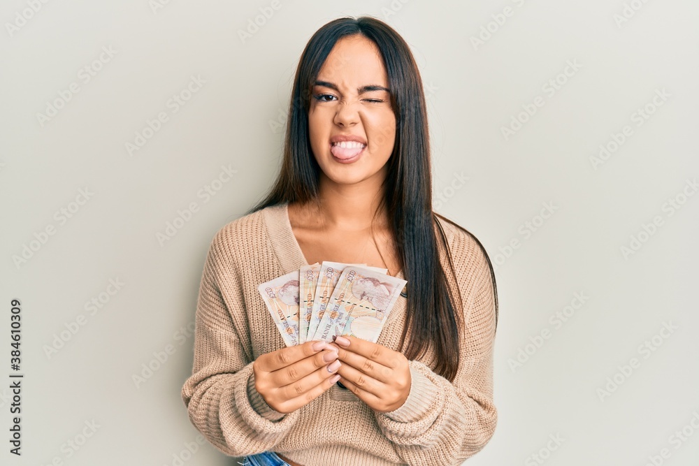 Young beautiful hispanic girl holding 10 united kingdom pounds banknotes sticking tongue out happy with funny expression. Young beautiful hispanic girl holding 10 united kingdom pounds banknotes sticking tongue out happy with funny expression.