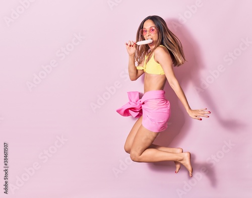 Young beautiful girl wearing bikini and sunglasses smiling happy. Jumping with smile on face holding lollipop over isolated pink background