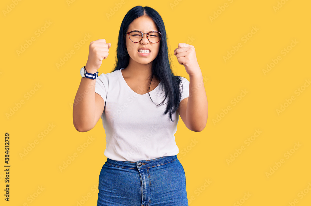 Young beautiful asian girl wearing casual clothes and glasses angry and mad raising fists frustrated and furious while shouting with anger. rage and aggressive concept. Young beautiful asian girl wearing casual clothes and glasses angry and mad raising fists frustrated and furious while shouting with anger. rage and aggressive concept.