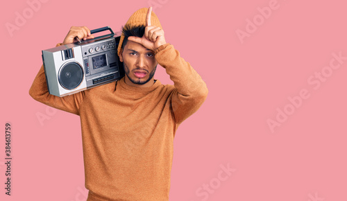 Handsome latin american young man holding boombox, listening to music making fun of people with fingers on forehead doing loser gesture mocking and insulting.