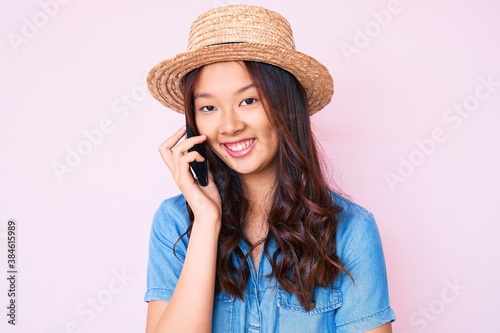 Young beautiful chinese girl wearing summer hat having conversation talking on the smartphone looking positive and happy standing and smiling with a confident smile showing teeth