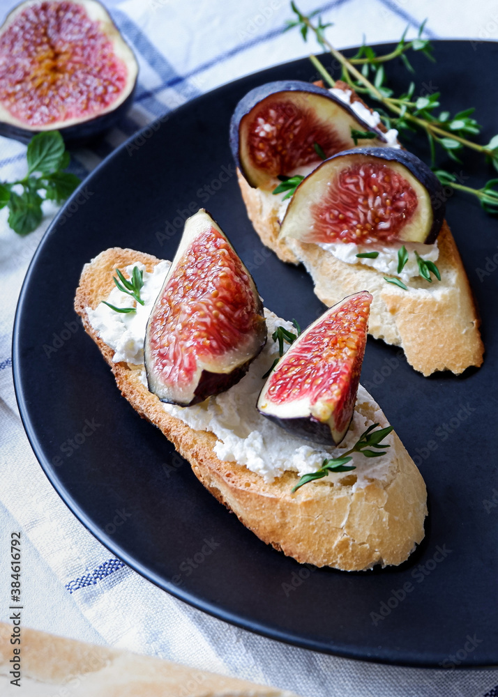 Two sandwiches with goat cheese, fig and thyme on fresh crispy bread served on a black plate on checked white and blue tablecloth