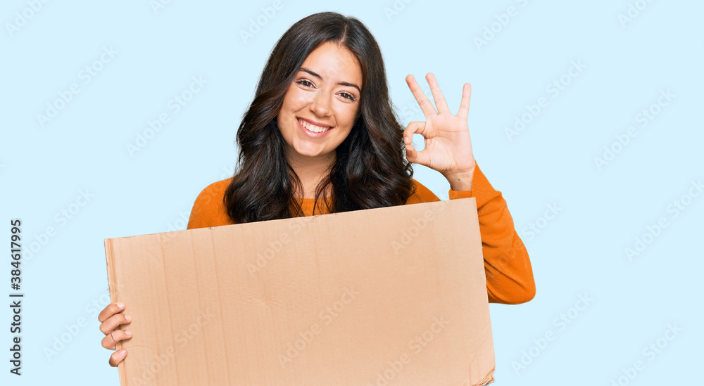 Beautiful brunette young woman holding cardboard blank empty banner doing ok sign with fingers, smiling friendly gesturing excellent symbol Beautiful brunette young woman holding cardboard blank empty banner doing ok sign with fingers, smiling friendly gesturing excellent symbol