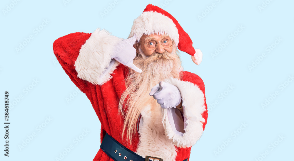 Old senior man with grey hair and long beard wearing traditional santa claus costume smiling doing talking on the telephone gesture and pointing to you. call me.