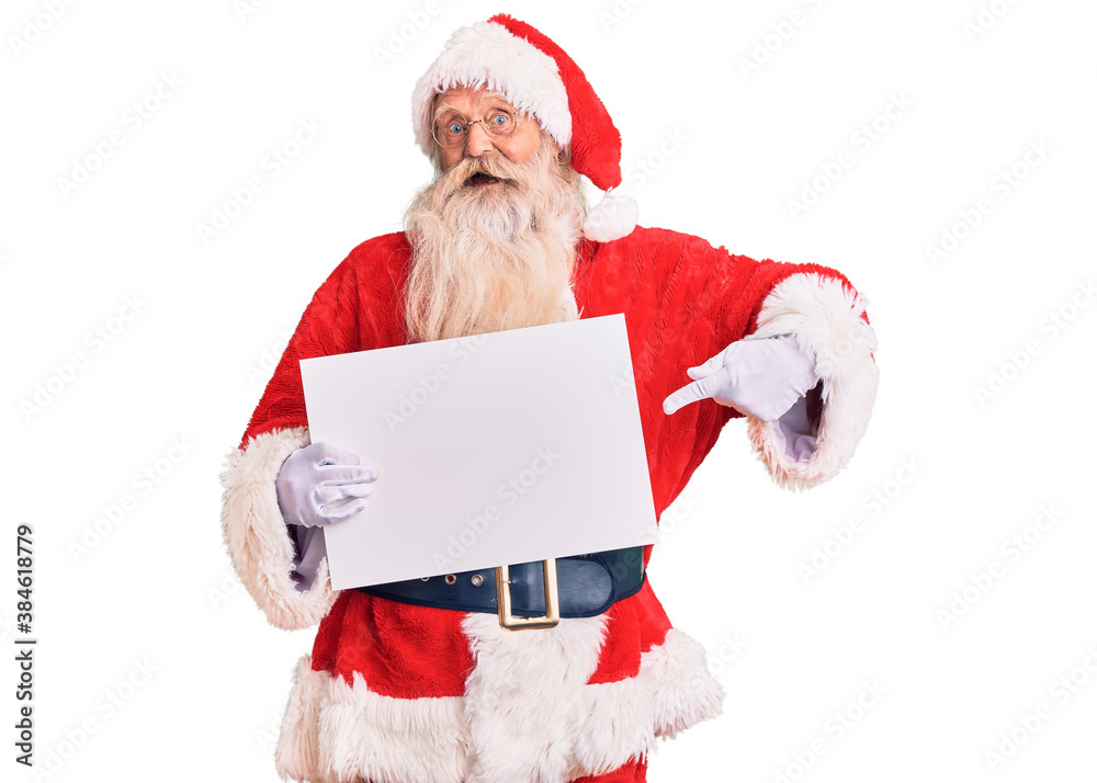 Naklejka premium Old senior man with grey hair and long beard wearing santa claus costume holding banner smiling happy pointing with hand and finger