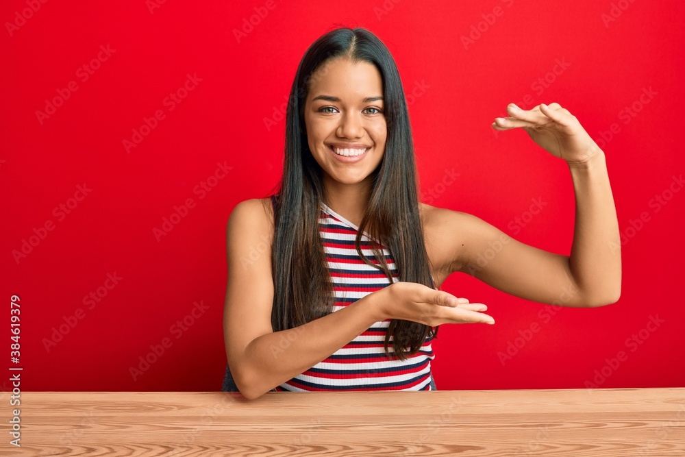 Beautiful hispanic woman wearing casual clothes sitting on the table gesturing with hands showing big and large size sign, measure symbol. smiling looking at the camera. measuring concept. Beautiful hispanic woman wearing casual clothes sitting on the table gesturing with hands showing big and large size sign, measure symbol. smiling looking at the camera. measuring concept.