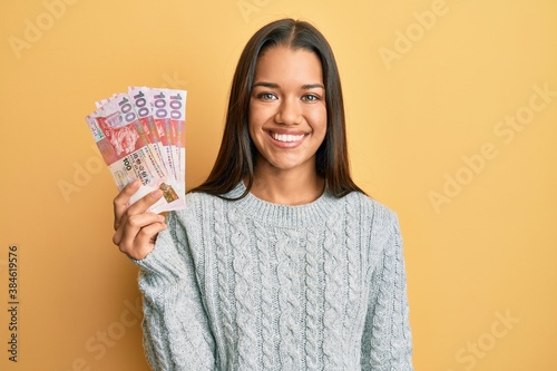 Beautiful hispanic woman holding 100 hong kong dollars banknotes looking positive and happy standing and smiling with a confident smile showing teeth