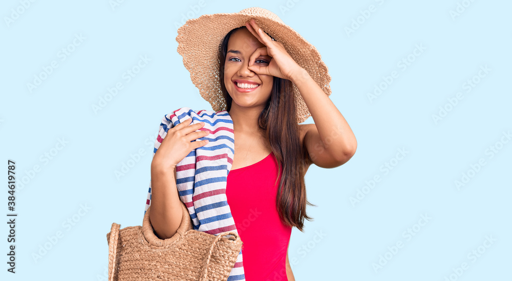 Young beautiful latin girl wearing swimwear and summer hat holding beach towel and bag smiling happy doing ok sign with hand on eye looking through fingers Young beautiful latin girl wearing swimwear and summer hat holding beach towel and bag smiling happy doing ok sign with hand on eye looking through fingers