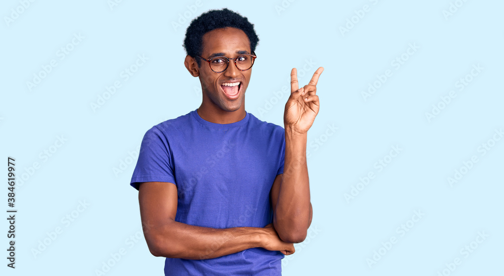 Naklejka premium African handsome man wearing casual clothes and glasses smiling with happy face winking at the camera doing victory sign. number two.