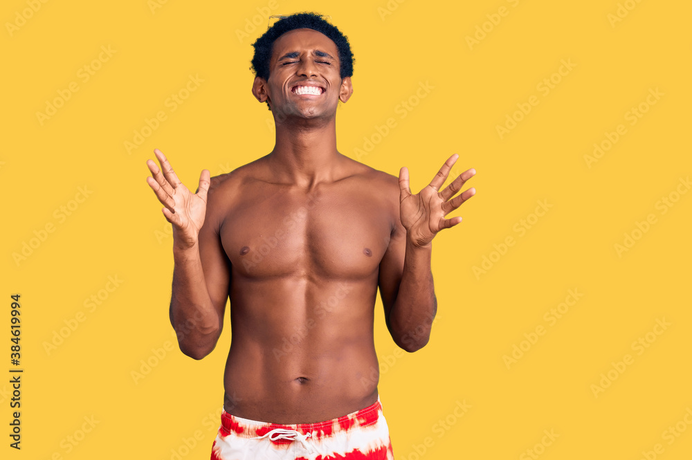 Fototapeta premium African handsome man wearing swimsuit and sunglasses celebrating mad and crazy for success with arms raised and closed eyes screaming excited. winner concept