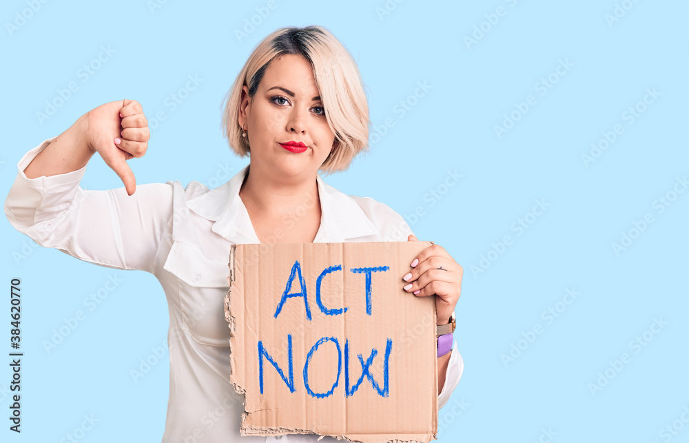 Young blonde plus size woman holding act now cardboard banner with angry face, negative sign showing dislike with thumbs down, rejection concept Young blonde plus size woman holding act now cardboard banner with angry face, negative sign showing dislike with thumbs down, rejection concept