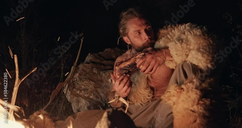 Primitive hunter gather wearing sheepskin squatting eating meat by the light of the flames of a fire at night tearing the meat from the bone with his teeth - wild caveman